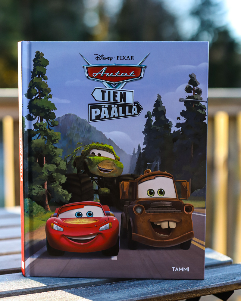Kuvassa on lastenkirja âDisney Pixar Autot. Tien pÀÀllĂ€â etukansi suoraan kohti katsojaa. Kannen kuvassa hymyilevĂ€t kirjan hahmot Salama McQueen ja hĂ€nen ystĂ€vĂ€nsĂ€ Martti. Kirja on pystyssĂ€ puisella pöydĂ€llĂ€ ulkona. Miljöö on kesĂ€inen.