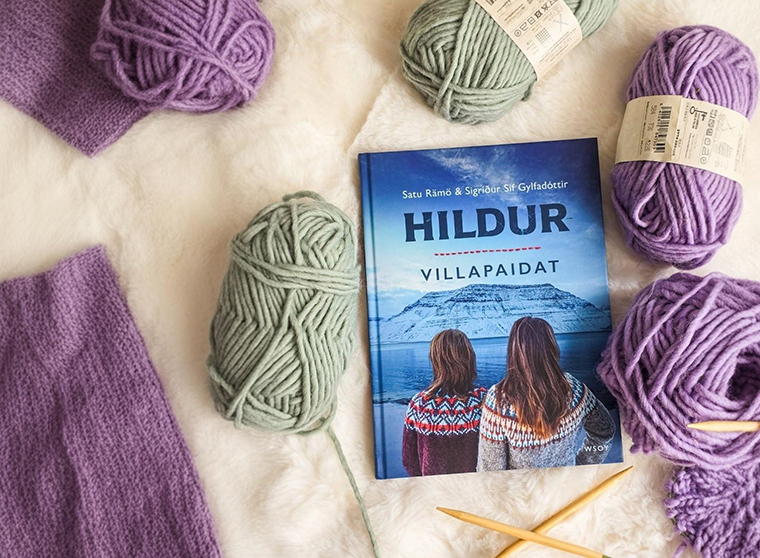 Kuvassa pehmeällä alustalla Hildur-villapaidat -kirja. Kirjan ympärillä on lankakeriä ja kutomapuikot sekä jo kudottua materiaalia.