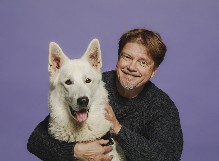 Kuva Timo Parvelasta halaamassa suurta, valkoista koiraa violetilla taustalla.a