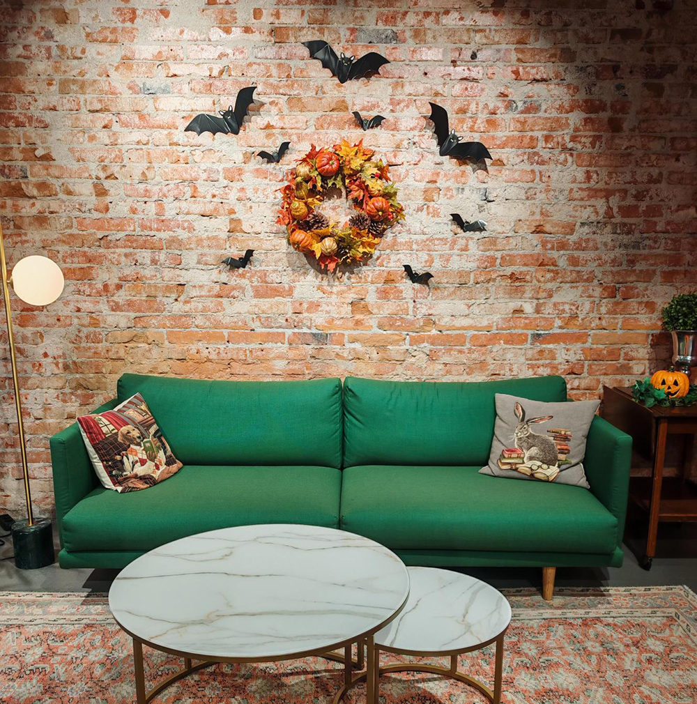 Kuvassa on vihreä sohva, jonka taakse tiiliseinään on kiinnitetty erilaisia koristeita kuten lepakoita ja syksyinen kranssi. 