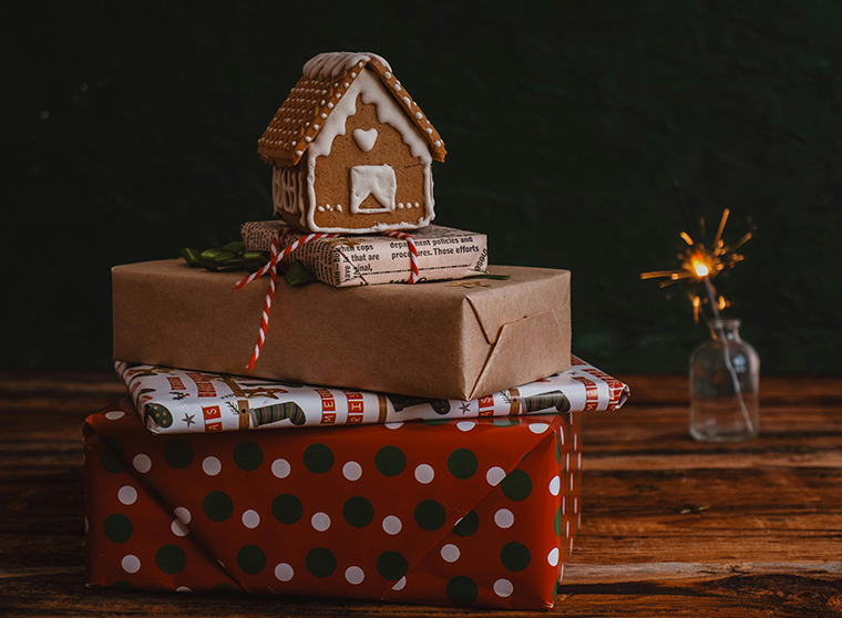 Kuvassa on punertavaksi petsatun lankkulattian päällä neljä joululahjaa pinossa. Alimpana pinossa oleva suurin paketti on kääritty pilkulliseen lahjapaperiin ja sen päällä on joulusukkakuvin koristeltuun lahjapaperiin kääritty paketti. Tämän päällä pinossa on ruskeaan paperiin kääritty paketti. Ylimpänä on sanomalehtipaperiin kääritty pieni paketti, ja sen päälle on asetettu valkoisella sokerikuorrutteella koristeltu piparkakkutalo. Kauempana, pakettipinon takana, näkyy palava tähtisadetikku. Kuva on tummasävyinen.