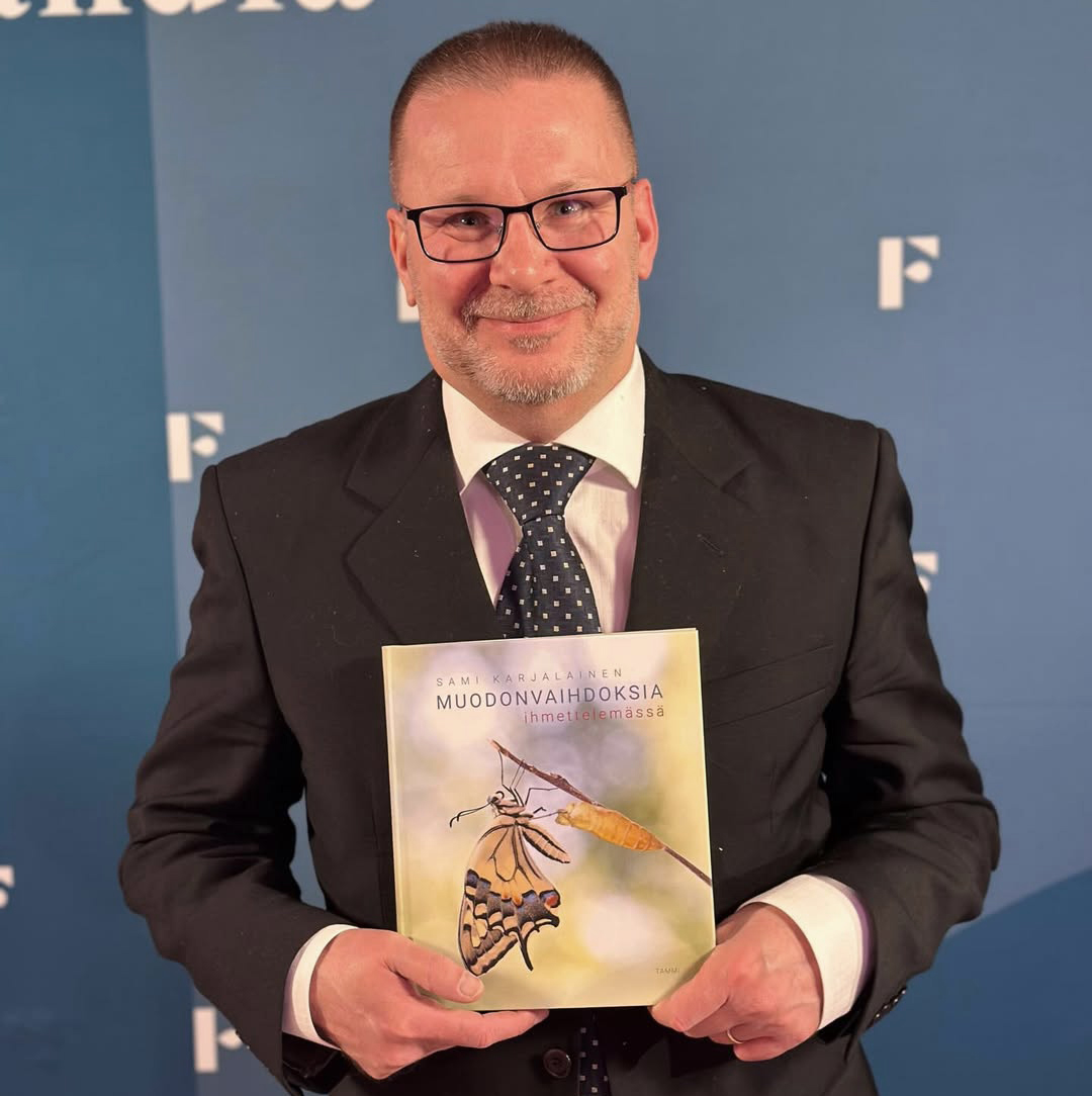 Sami Karjalainen seisoo puku päällä, katsoo kameraan hymyillen ja pitelee kirjaansa Muo­don­vaih­dok­sia ih­met­te­le­mäs­sä.