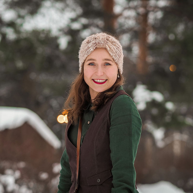 Kuvassa on Lotta-Sofia Saahko kuvattuna vyötäröstä ylöspäin. Hän seisoo ulkona talvisessa metsässä ja on kääntynyt leveästi hymyillen katsomaan suoraan kohti katselijaa. Hänellä on yllään tummanvihreä paita, tummanruskea liivi ja päässään vaaleanruskea villapanta.