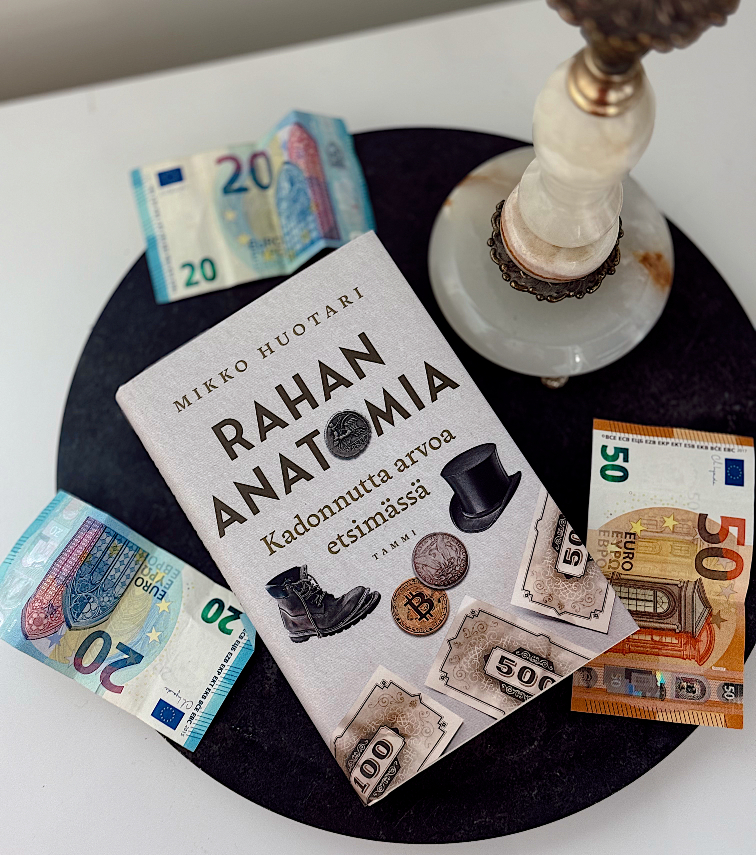 Kuvassa makaa mustalla pyöreällä pöydällä kirja ”Rahan anatomia”. Kirja on kuvattu suoraan ylhäältäpäin. Kirjan ympärillä pöydällä on kaksi kahdenkymmenen euron seteliä ja yksi viidenkymmenen euron seteli. Pöydällä on myös valkoinen marmorinen kynttiläjalka.