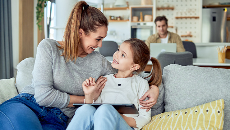 Vrouw en kind op de bank met een tablet en man achter een tafel met een laptop