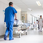 Zorgmedewerker in blauwe werkkleding loopt door een ziekenhuisgang met bedden en personeel op de achtergrond
