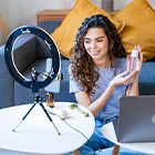 Vrouw zit aan tafel met laptop en ringlamp en toont een productflesje aan de camera
