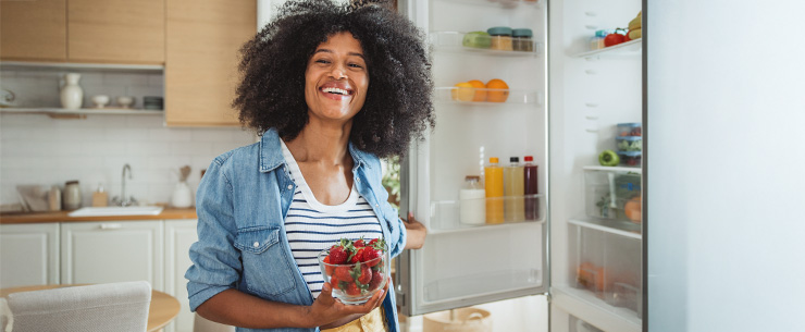 Vrouw staat voor een open koelkast en houdt een schaaltje aardbeien vast in een keukenomgeving