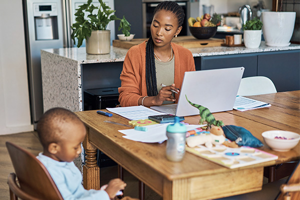 Vrouw werkt aan een laptop aan de keukentafel terwijl een jong kind naast haar zit.