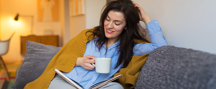 Persoon zit op een bank met een kopje en een tijdschrift in een woonruimte.