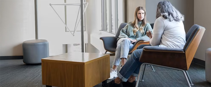 Twee personen zitten tegenover elkaar in gesprek in een wachtruimte met stoelen en een tafel.