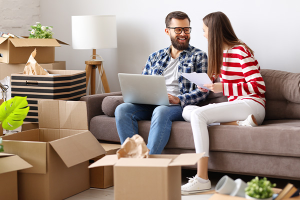Twee personen zitten op een bank tussen verhuisdozen en bekijken documenten en een laptop.