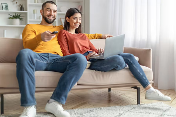 Man en vrouw op een bank, waarbij de man een afstandsbediening vasthoudt en de vrouw een laptop gebruikt