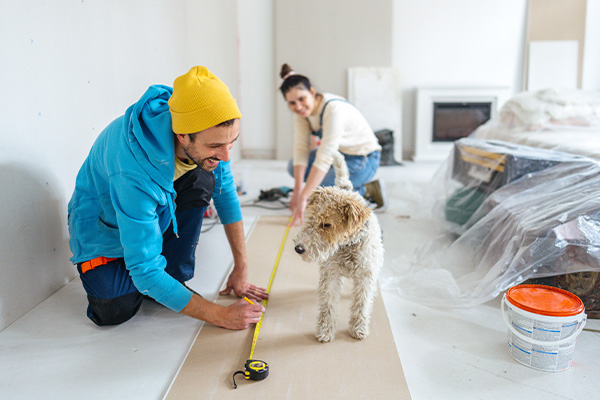 Man meet plank op vloer terwijl vrouw knielt; hond staat ertussen in ruimte met afdekfolie