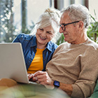 Een oudere vrouw en man zitten naast elkaar en kijken glimlachend naar een laptop in een lichte woonkamer