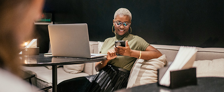 Vrouw zit met smartphone op een bank naast een open laptop.