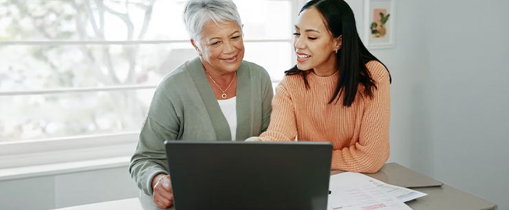 Twee vrouwen bekijken samen een laptop aan tafel met documenten ernaast.