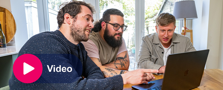 Drie mannen kijken samen naar een laptop aan tafel, met links een knop met de tekst Video.