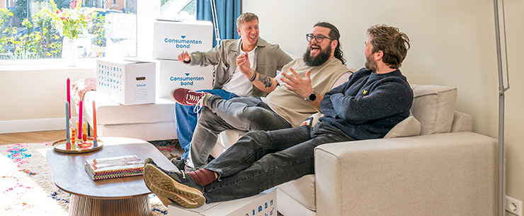 Drie mannen lopen met verhuisdozen naar een woning in een straat met geparkeerde auto’s.