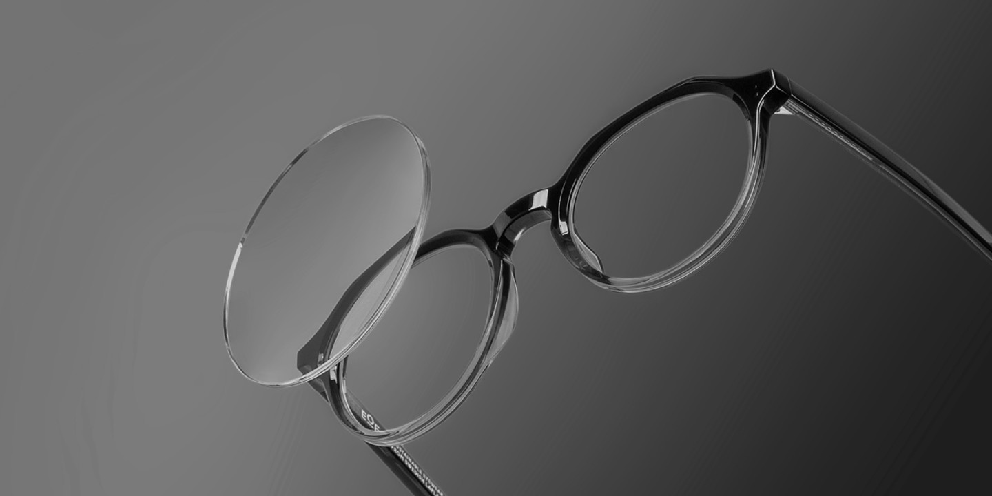 Schwarz-weiß-aufnahme einer Brille mit rundem Gestell; ein Brillenglas ist aus dem Rahmen herausgelöst und schwebt leicht davor.