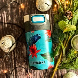 Turquoise Kambukka insulated travel mug with tropical bird and red flower design on wooden surface with festive bells and pine