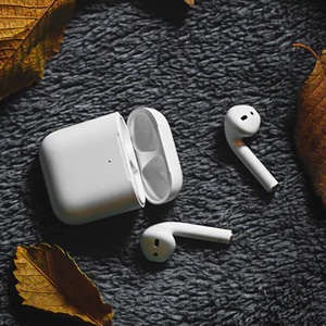 White true wireless earbuds with open charging case on dark textured fabric, surrounded by autumn leaves