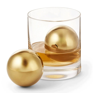 Clear glass tumbler of whisky with two gold spherical chilling stones, one inside glass and one beside on white background