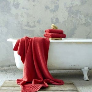 White clawfoot bathtub in a rustic bathroom with red towels draped and stacked and a wooden bath brush on the rim