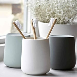 Three ceramic toothbrush cups (white, grey, green) holding bamboo toothbrushes on a windowsill with plant