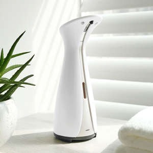 White automatic foam soap dispenser on bathroom counter beside a potted plant and towel, with window blinds in the background