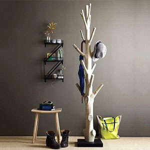 Tree-shaped wooden coat rack in a modern hallway holding a hat and scarf, beside boots, stool, wall shelves and a yellow tote bag