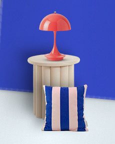 Runde, beige Tisch mit roter Lampe und gestreiftem Kissen in Blau und Rosa vor blauem Hintergrund.