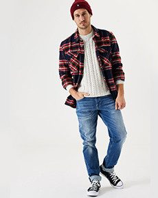 Person in lässiger Kleidung mit einem karierten Hemd, einem weißen Pullover, Jeans und Sneakers sowie einer roten Mütze.