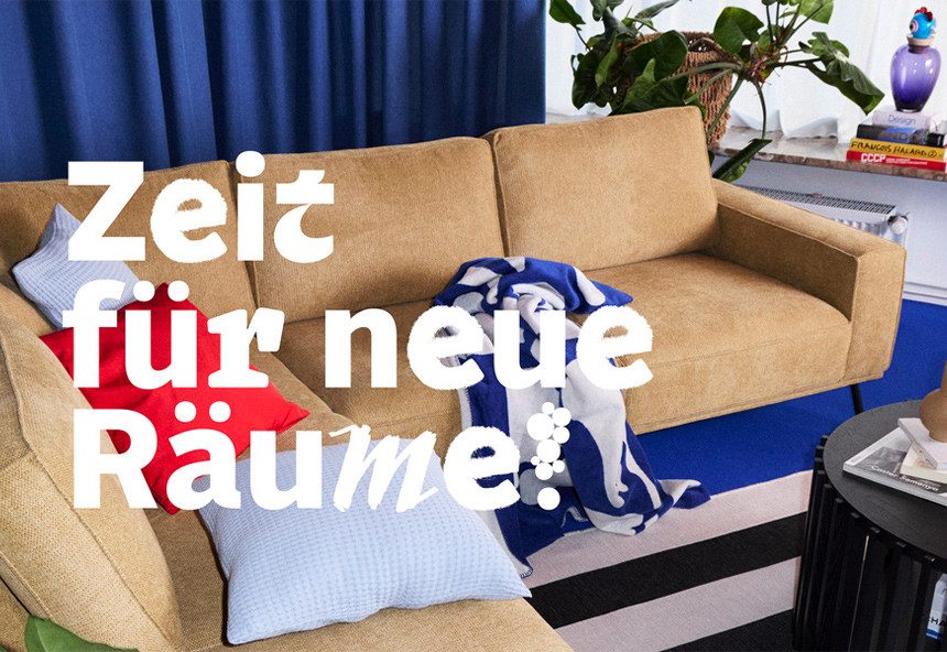 Text mit „Zeit für neue Räume!“ in großen, weißen Buchstaben, vor einem beigen Sofa mit bunten Kissen und einer blauen Decke in einem wohnlichen Raum.