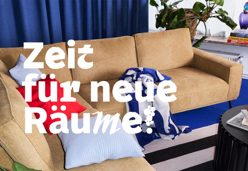 Beige Couch mit bunten Kissen vor blauem Teppich und schwarzem Couchtisch. Text: „Zeit für neue Räume!“