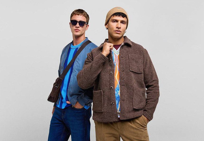 Zwei Männer stehen vor einem hellen Hintergrund. Der linke trägt eine blaue Jacke, einen blauen Pullover und eine Umhängetasche. Der rechte trägt eine braune Jacke, einen bunten Pullover und eine beige Mütze.