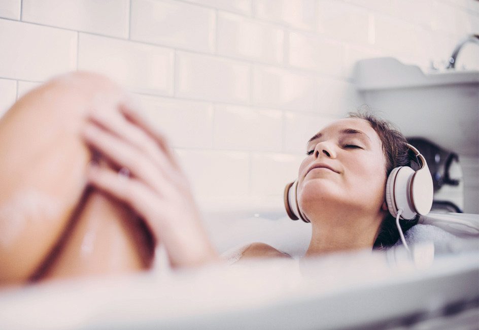 Frau mit geschlossenen Augen und Kopfhörern liegt genussvoll in der Badewanne