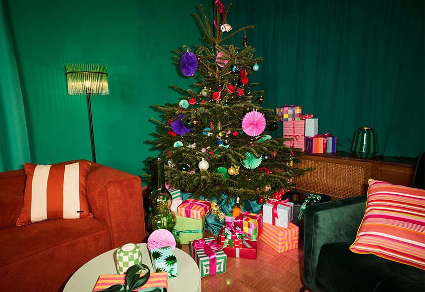 Foto von einem gemütlichen Wohnzimmer mit einem orangenen Sofa, bunten Kissen und einem geschmückten Weihnachtsbaum mit Geschenken darunter. Neben dem Baum steht ein kleiner runder Tisch mit einer roten Lampe.