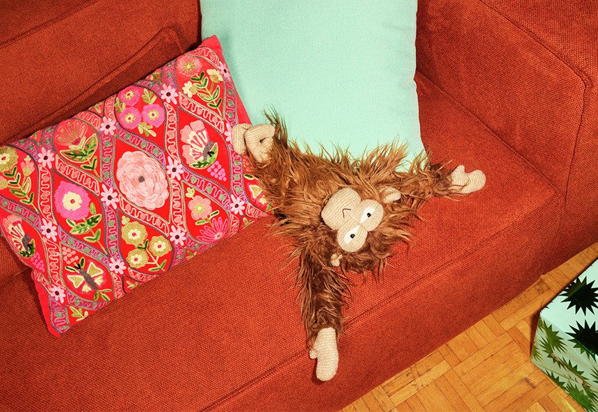 Ein weiches Kuscheltier in Form eines Affen liegt auf einem roten Sofa zwischen einem bunten Kissen und einem hellblauen Kissen.