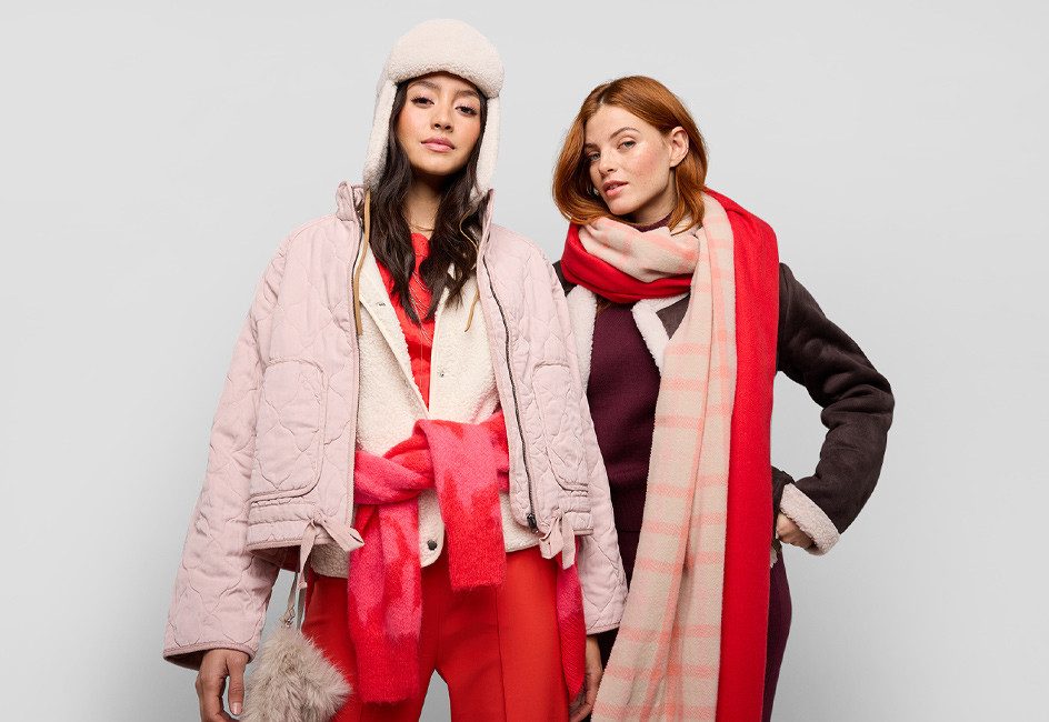 Zwei Frauen in stylischer Winterkleidung wie auffälliger Mütze und voluminösem Schal stehend vor Grau