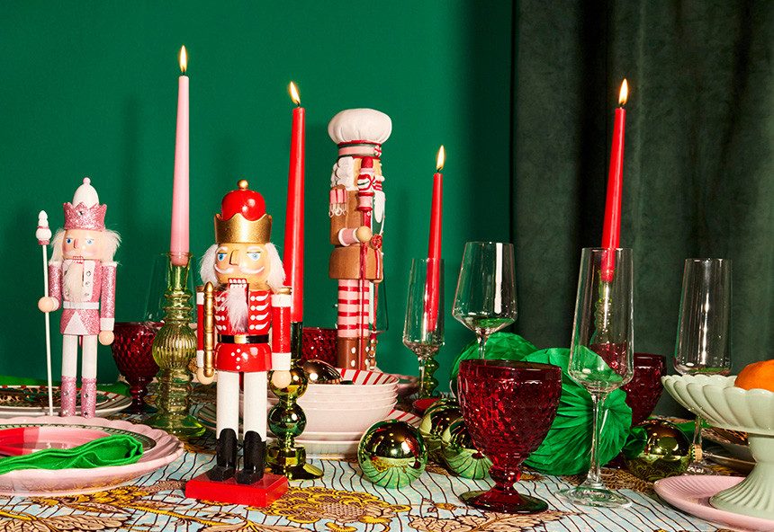 Auf dem Bild sieht man einen festlich gedeckten Tisch mit drei Nussknacker-Figuren und brennenden Kerzen. Der Hintergrund ist grün und links und rechts stehen verschiedene Gläser und Dekorationen.