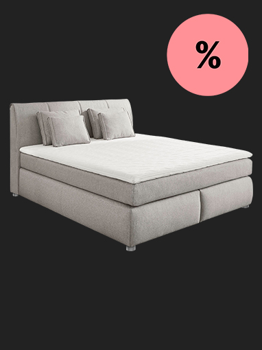 Graues Boxspringbett mit Kissen, rosa Prozentzeichen deutet auf Preisnachlass hin.