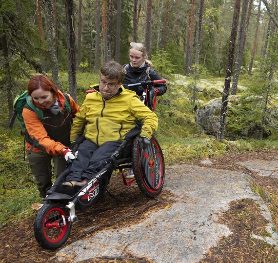 Kuvassa kaksi henkilöä työntää kolmatta maastopyörätuolissa ulkona metsässä.