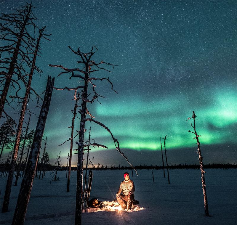 Mies istumassa nuotion ääressä lumihangen keskellä. Taivaalla näkyy kirkkaat revontulet.