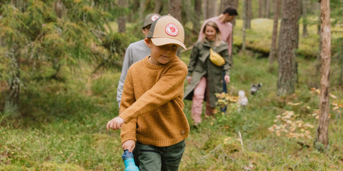 Lapsi kävelemässä iloisena metsässä. Hänellä on kädessään juomapullo. Taustalla näkyy muu perhe.