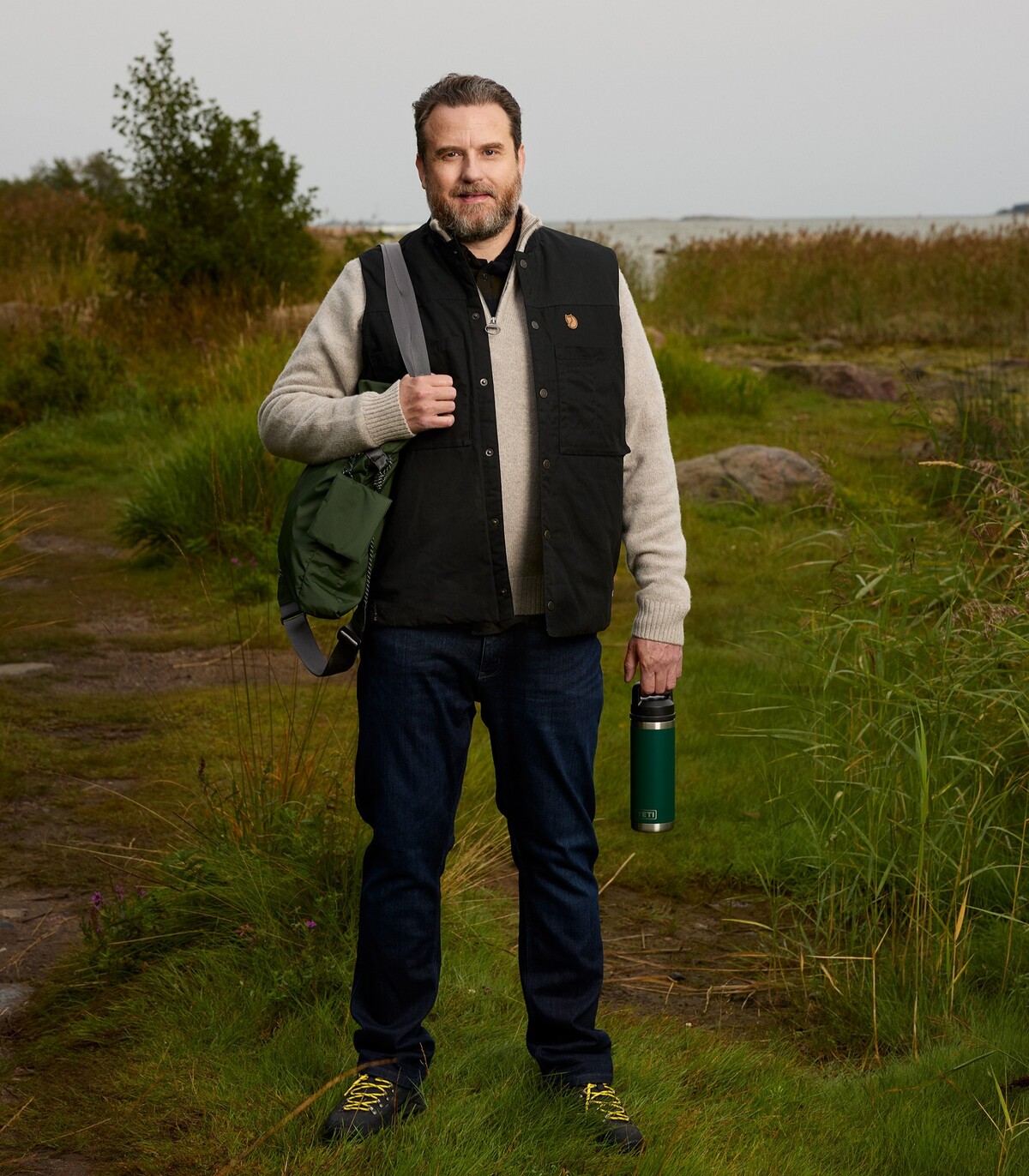 Vanhempi mies kuvattuna nurmikolla. Hän katsoo hymyillen kameraan. Hänellä on toisella olallaan reppu ja toisessa kädessä juomapullo