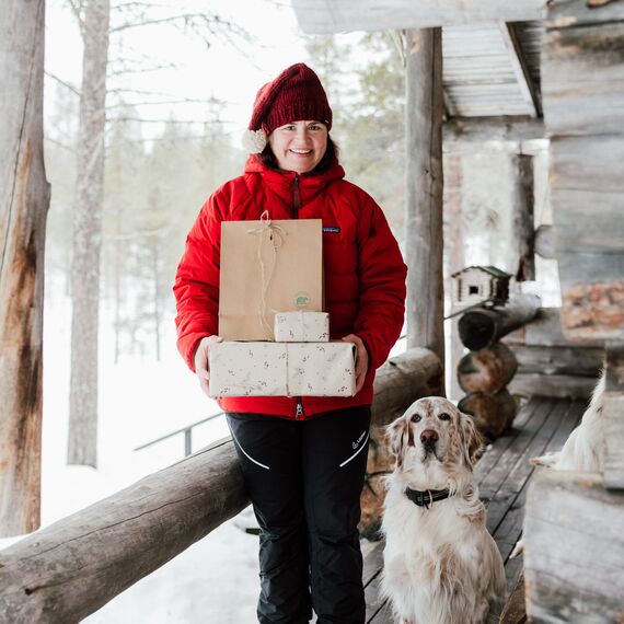 Nainen seisomassa kuistilla. Hänellä on käsissään joulupaketteja. Hänen vierellään on koira.