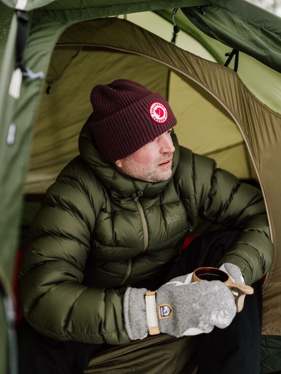 Mies katsomassa ulos teltasta. H&auml;nell&auml; on talvivaatteet p&auml;&auml;ll&auml;&auml;n ja muki k&auml;dess&auml;