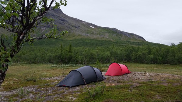 Hillebergin Niak-teltta on pystytetty rinteeseen. On auringonlastku ja auringons&auml;teet korostavat taustalla olevan vuoristomaiseman &auml;&auml;riviivoja. Pilvet ovat dramaattisia.