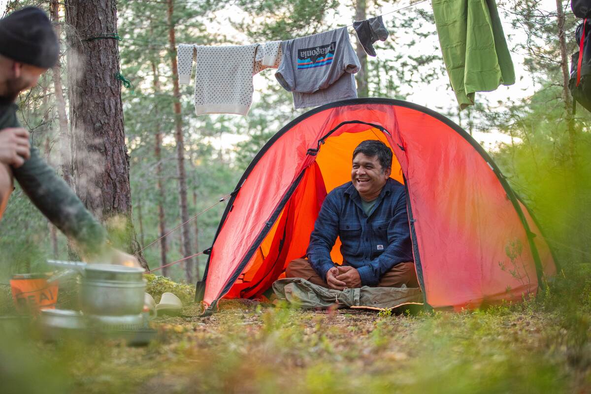 hillebergin Nallo 2 GT j&auml;rvimaisemassa. Kaksi ihmist&auml; istuu teltan edustalla. Teltta on pystytetty pienelle niemelle ja kuvajainen heijastuu j&auml;rveen.